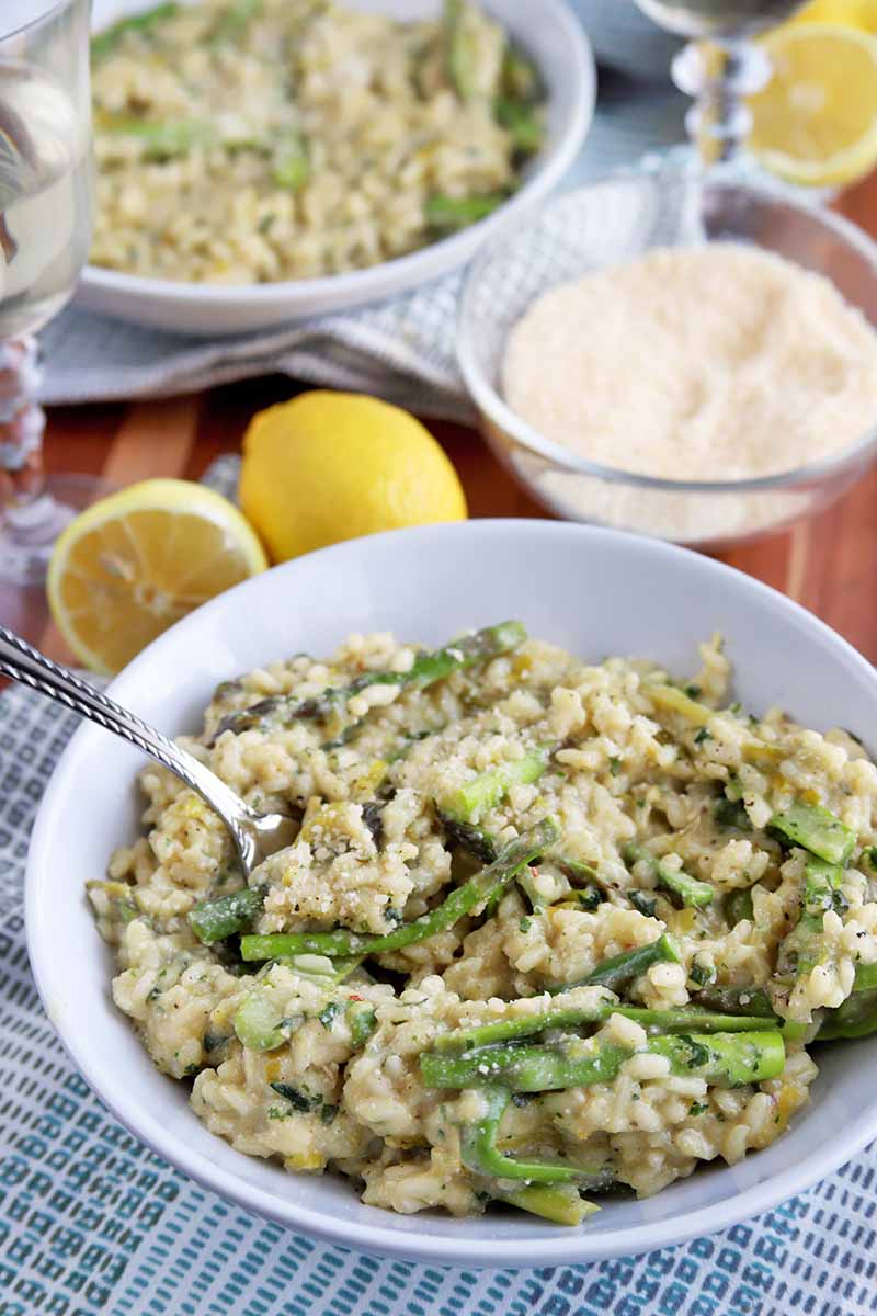 Vertical image of two white bowls of homemade asparagus risotto, on blue and white checkered cloth napkins on a brown wood table, with two glasses of white wine, a small glass bowl of grated cheese, whole and halved lemons, and forks.