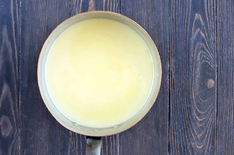 Overhead horizontal image of a saucepan filled with a yellow pudding mixture, on a dark brown wood table.