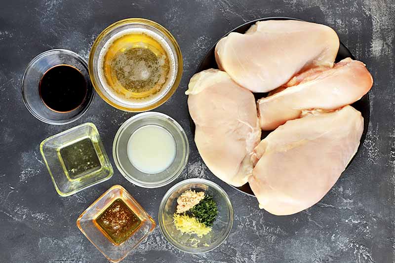 Overhead shot of two small square and four small round glass prep bowls of oil, soy sauce, beer, minced garlic, honey, and fresh herbs, beside a black plate of four raw chicken breasts without bone or skin, on a gray and white background.