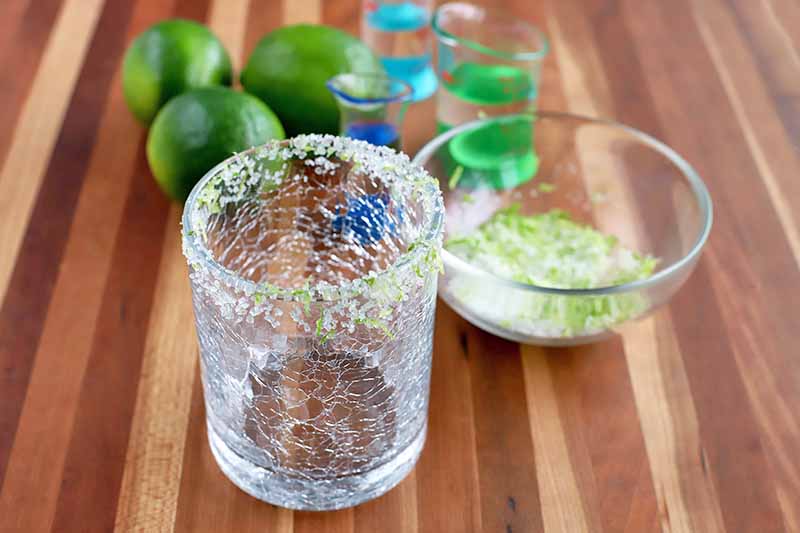 Slightly oblique head-on horizontal shot of a rocks glass with a salt and citrus zest rim, a small glass dish of the rimming mixture, three limes, and two measuring beakers of liquor, on a brown wood surface with vertical stripes.