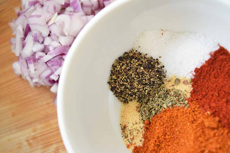 Overhead closely cropped image of a small pile of chopped purple onion to the left, and a white bowl with piles of several different types of spices, salt, and ground black pepper to the right, on a wooden cutting board.