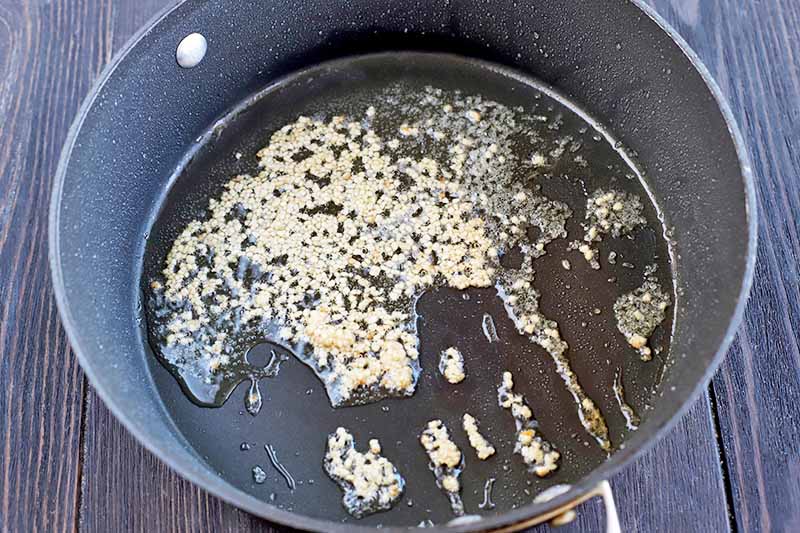 A large nonstick frying pan with high walls, with minced garlic sauteeing in oil at the bottom, on a dark brown wood surface.