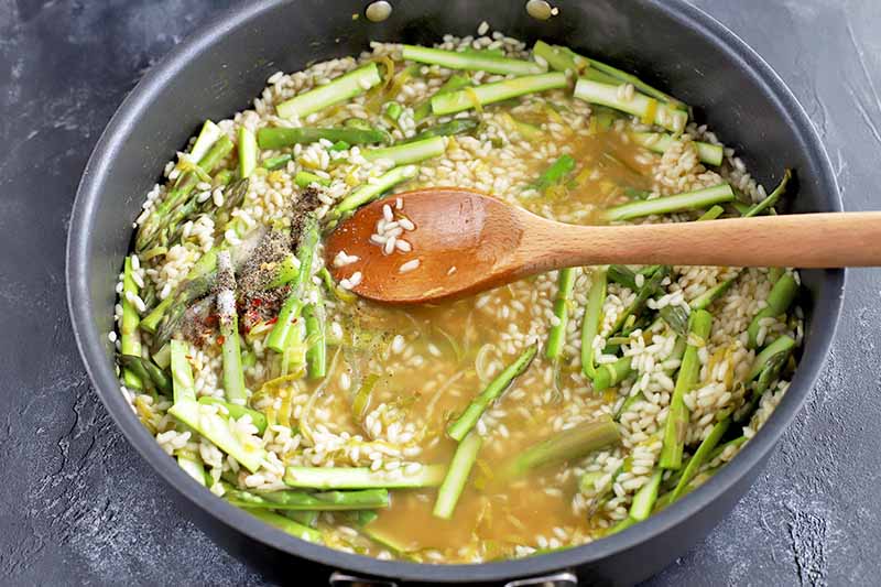 Horizontal image of a large frying pan of broth, medium-grain white rice, asparagus, and leeks, being stirred with a wooden spoon, on a gray slate surface.