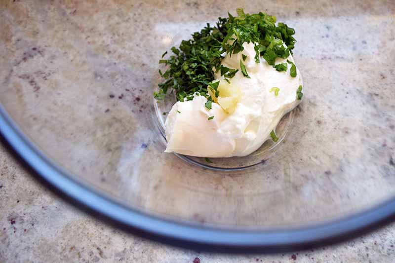 Overhead slightly oblique closeup closely cropped image of a glass bowl with a dollop of mayonnaise and chopped fresh green herbs at the bottom.