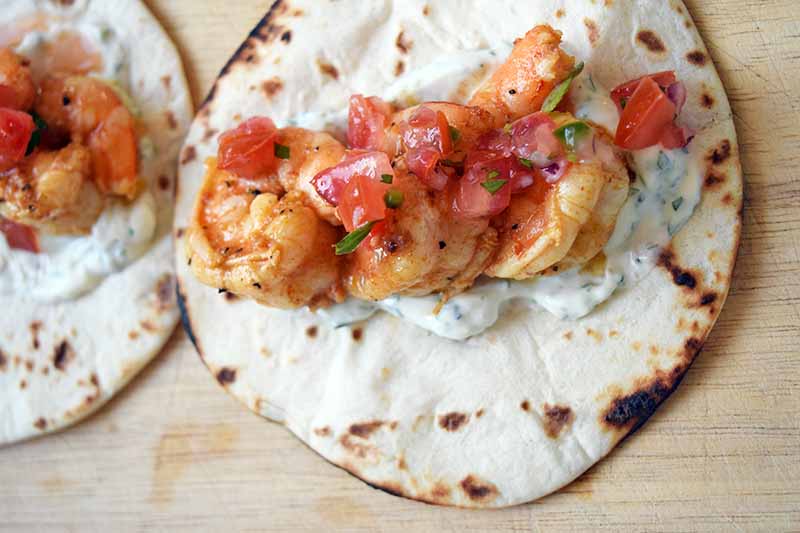 Overhead horizontal image of a toasted flour tortilla with three cooked shrimp on top of a smear of herb crema, with fresh pico de gallo salsa on top, and another identical taco to the left, on a wood cutting board.