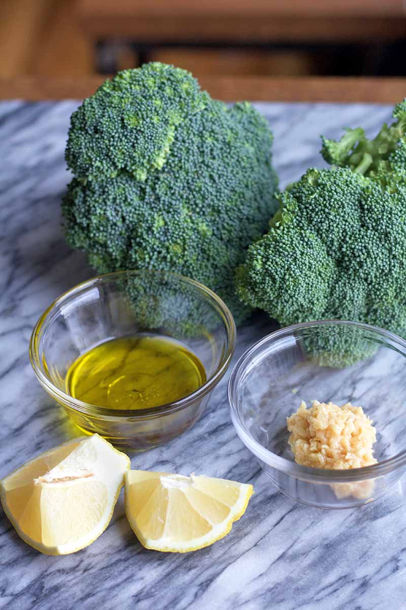 Vertical image of a marble surface topped with two heads of green broccoli, two small glass bowls of olive oil and minced garlic, and two lemon wedges, with brown kitchen cabinets in soft focus in the background.
