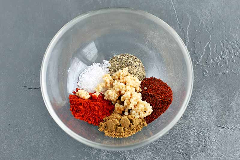 Overhead horizontal shot of a glass bowl filled with various spices and minced garlic in separate piles, about to be stirred together, on a gray surface.