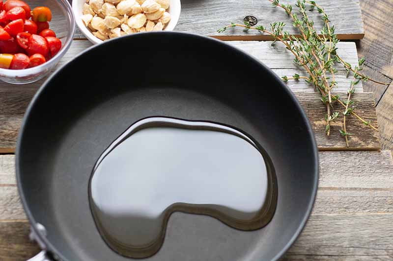 Overhead horizontal image of olive oil in a nonstick frying pan, with small glass bowls of halved red cherry tomatoes and blanched Marcona almonds, with sprigs of fresh thyme to the right, on an unfinished weathered wood surface.