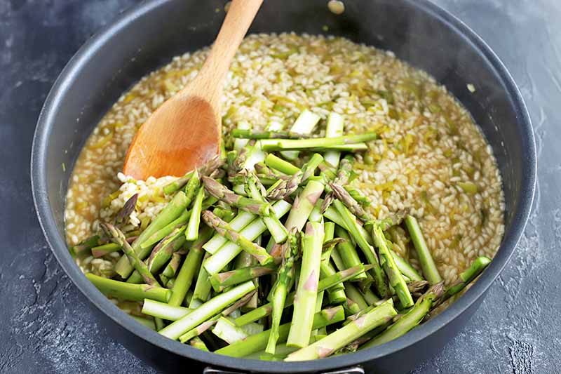 A wooden spoon stirs sliced fresh asparagus into a large frying pan of rice and broth, on a gray slate surface.