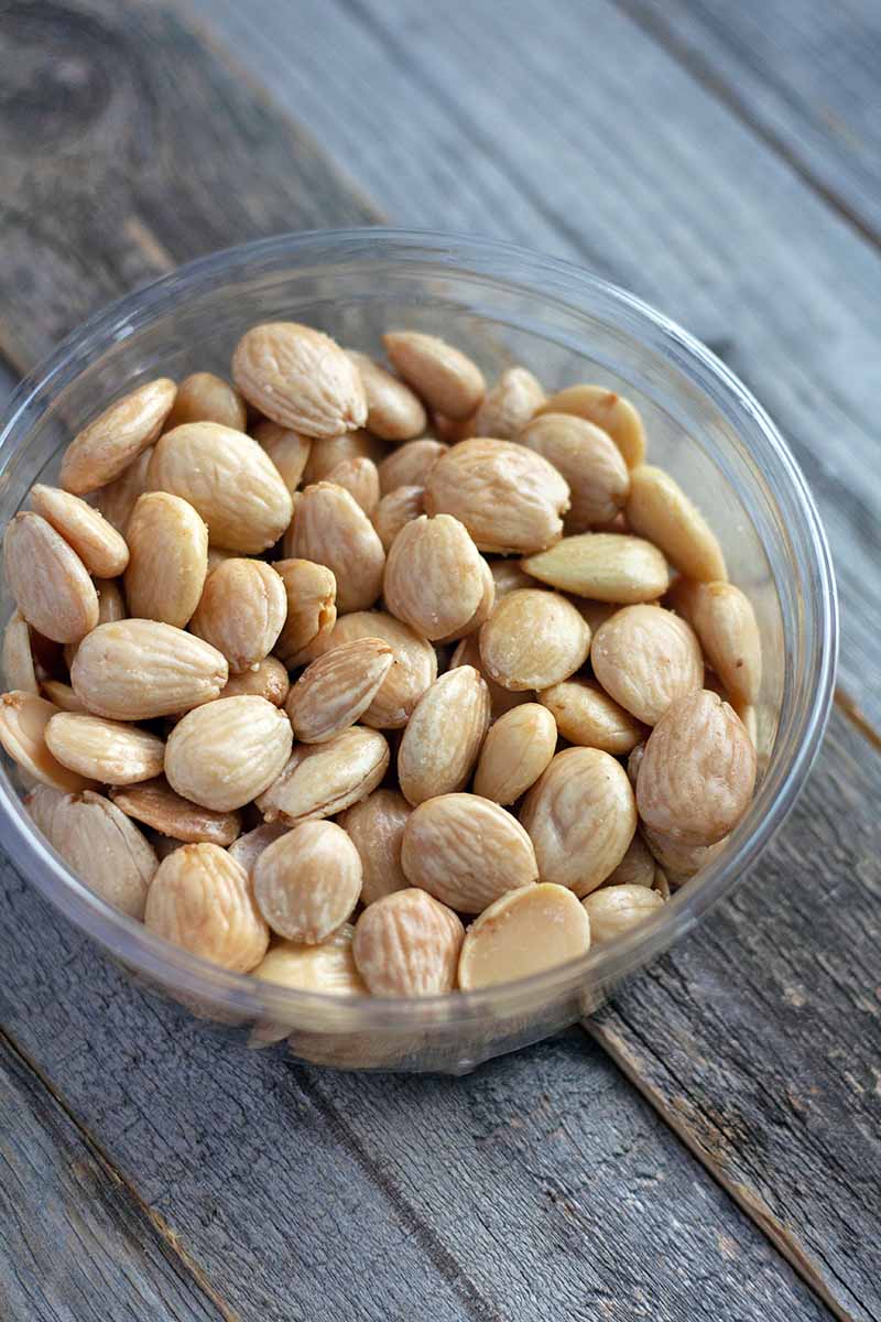Vertical overhead image of a small glass bowl of blanched whole Marcona almonds, on an unfinished wood surface.