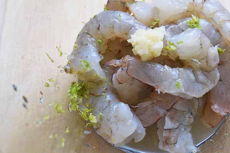 Closely cropped closeup horizontal image of raw shelled and deveined shrimp being sprinkled with minced garlic and lime zest, ona a beige wood background visible through a clear glass mixing bowl.