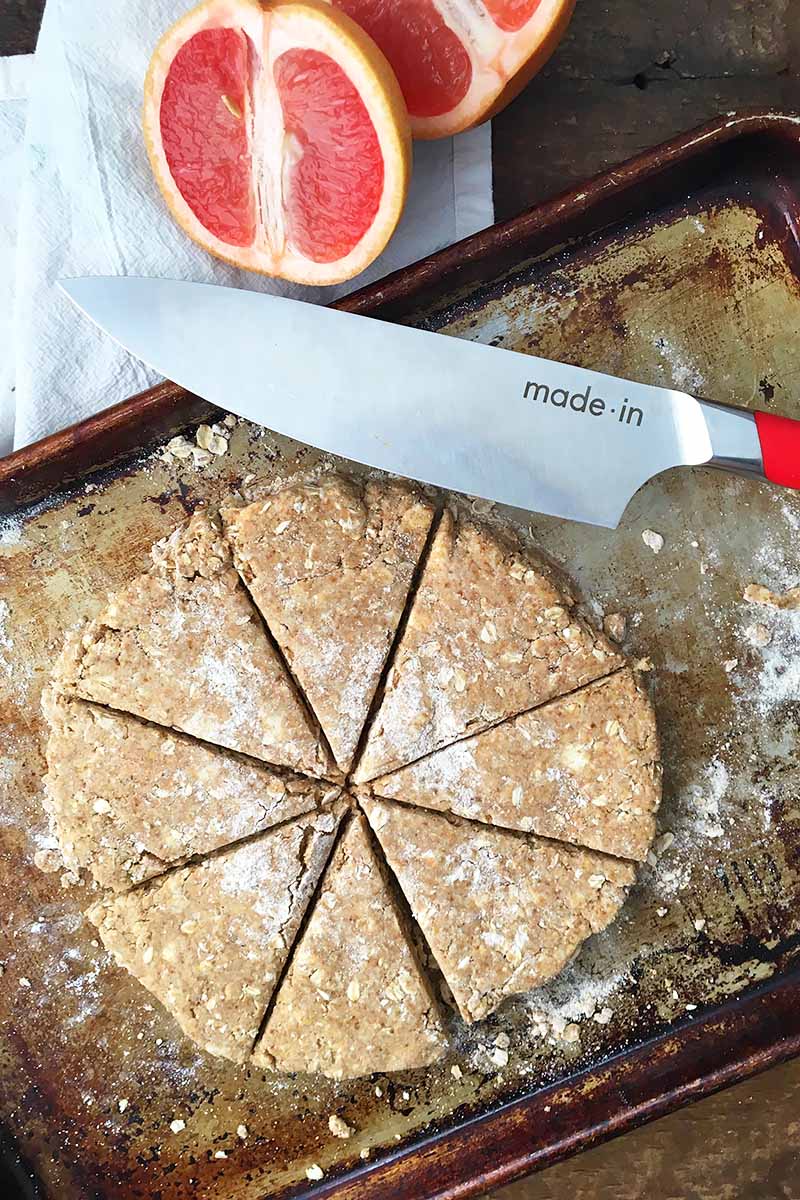 Vertical top-down image of a Made In knife next to portioned pieces of unbaked dough on lightly flour-dusted sheet pans, next to sliced grapefruit.