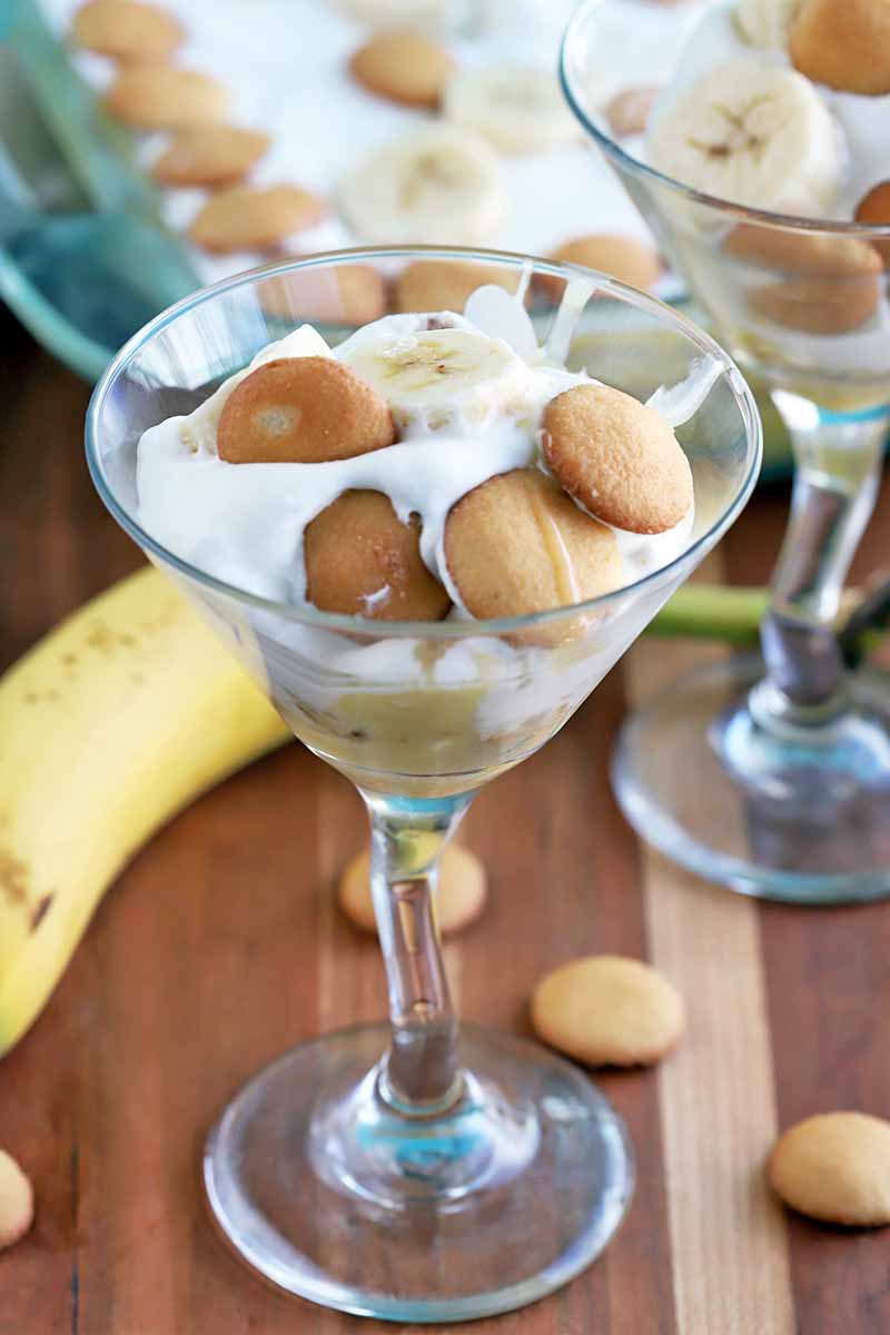 Vertical image of a martini glass of banana pudding with another identical glass and a square baking dish of the dessert in the background, on a striped brown and beige wood surface with scattered cookies and a yellow banana.