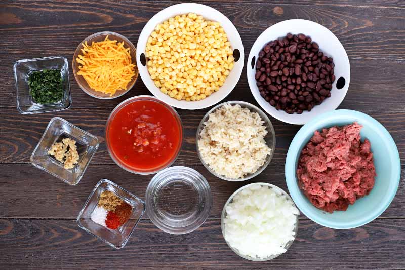 Overhead shot of round and square glass and ceramic bowls of various sizes, filled with a variety of ingredients required to make a Tex-Mex skillet dish including chopped fresh herb, minced garlic, spices, shredded cheese, salsa, corn, rice, onion, black beans, and ground beef, on a dark brown wood table.