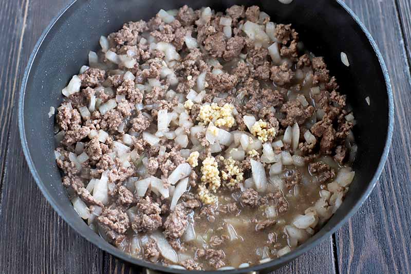 Horizontal image of a large frying pan of browned ground beef, sauteed onion, and raw minced garlic, on a dark brown wood background.