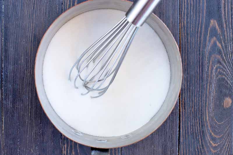 Overhead horizontal shot of a wire whisk stirring a white sugar and milk mixture in a metal saucepan, on a dark brown wood surface.