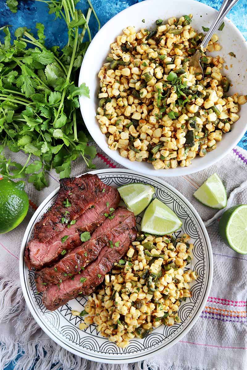 Vertical top-down image of a plate of corn and steak and a bowl of corn next to cilantro and limes.