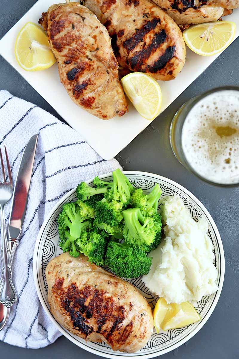 Vertical overhead shot of a white serving platter and a matching plate of grilled chicken with lemon wedges, with steamed broccoli and mashed potatoes, with a mug of beer to the right and a striped white cloth napkin topped with a knife and fork to the left, on a gray slate surface.