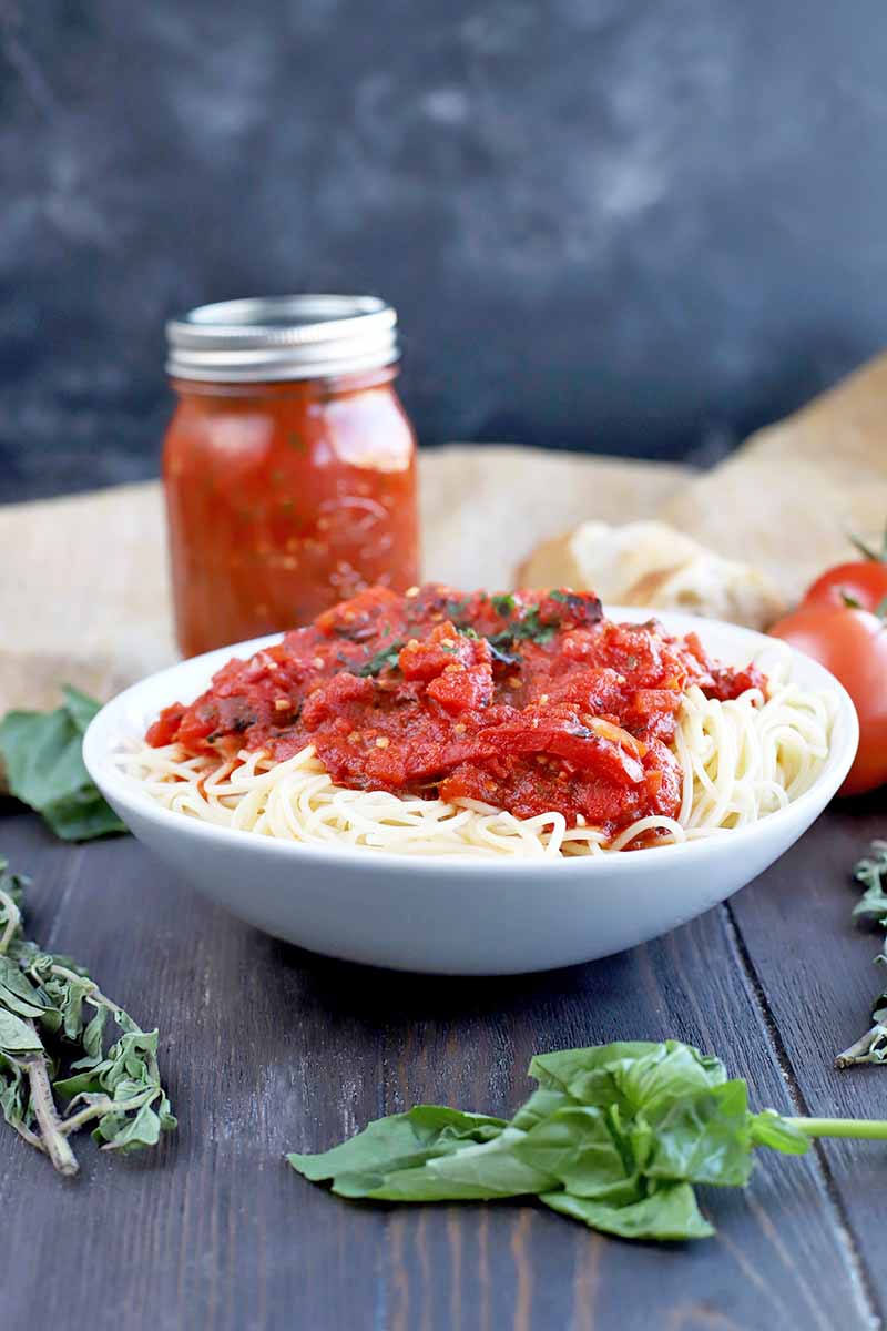 Vertical image of a white shallow bowl of spaghetti topped with chunky red tomato sauce, garnished with chopped herbs, with a jar of the same in soft focus in the background, on a dark brown wood surface with a folded piece of light brown burlap and sprigs of fresh basil and oregano, against a mottled gray backdrop.