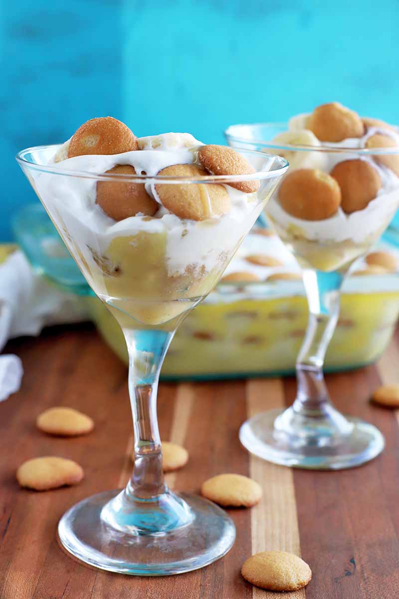 Vertical image of two martini glasses of pudding with whipped cream, banana slices, and miniature vanilla wafers, with more of the cookies scattered on a brown wood surface, with a glass dish of the dessert and a white cloth in the background, against a mottled blue backdrop.