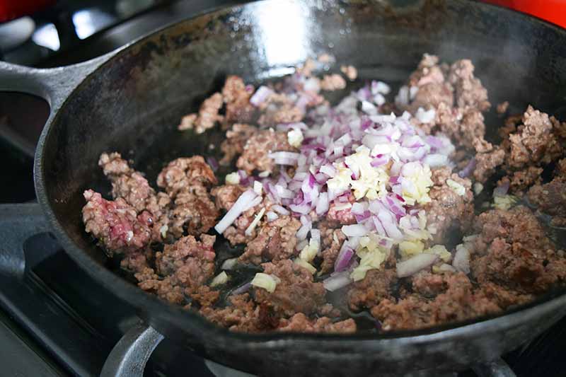 Horizontal image of browned ground beef with a pile of raw chopped red onion and minced raw garlic on top, on a large cast iron frying pan, on a gas stove with black metal grates.