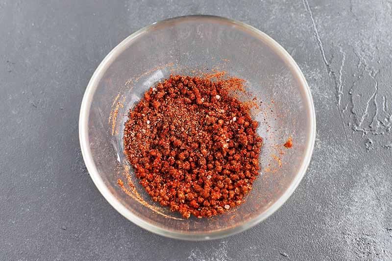 Overhead horizontal image of a red spice and garlic mixture in a glass bowl, on a gray surface.