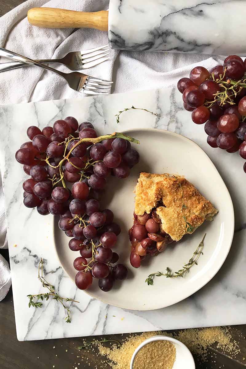 Vertical top-down image of a slice of grape pie on a white plate with more fresh fruit and fresh thyme leaves next to forks, sugar, and a rolling pin.