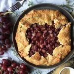 Horizontal image of a whole baked galette on a marble board next to whole grapes thyme leaves, sugar, and a metal fork.