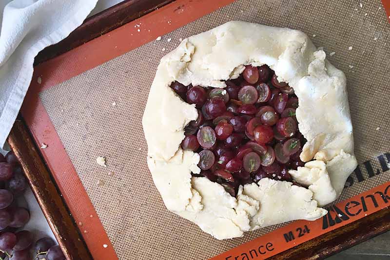 Horizontal image of an unbaked open-faced grape pie on a sheet pan lined with a silicone mat.