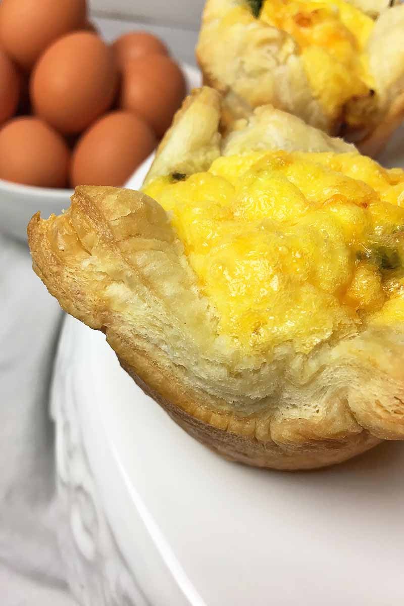 Vertical close-up image of one breakfast tart with puff pastry crust on a white stand, and eggs in a bowl in a background.