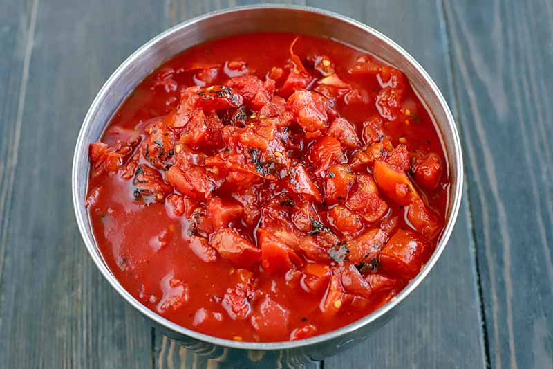 Horizontal overhead image of a stainless steel bowl of fire-roasted canned tomatoes in their juices, on a dark brown wood surface.