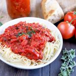 Horizontal image of a white shallow bowl of cooked spaghetti topped with marinara, on a brown wood table with tomatoes on the vine, a piece of baguette, a jar of sauce, and sprigs of basil and oregano, with a piece of burlap in the background.