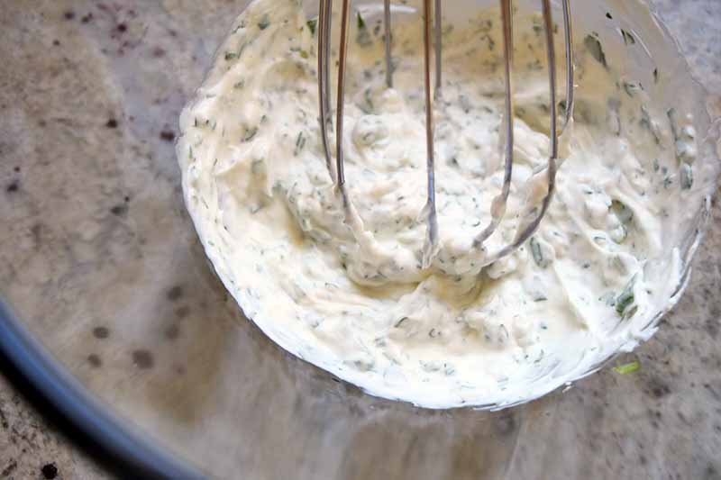 Overhead closely cropped closeup image of a wire whisk stirring a sour cream and herb mixture in a large glass mixing bowl, on a speckled beige and brown countertop.