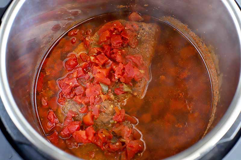 Overhead horizontal image of a stainless steel slow cooker insert filled with meat, cooking liquid, and stewed canned tomatoes.