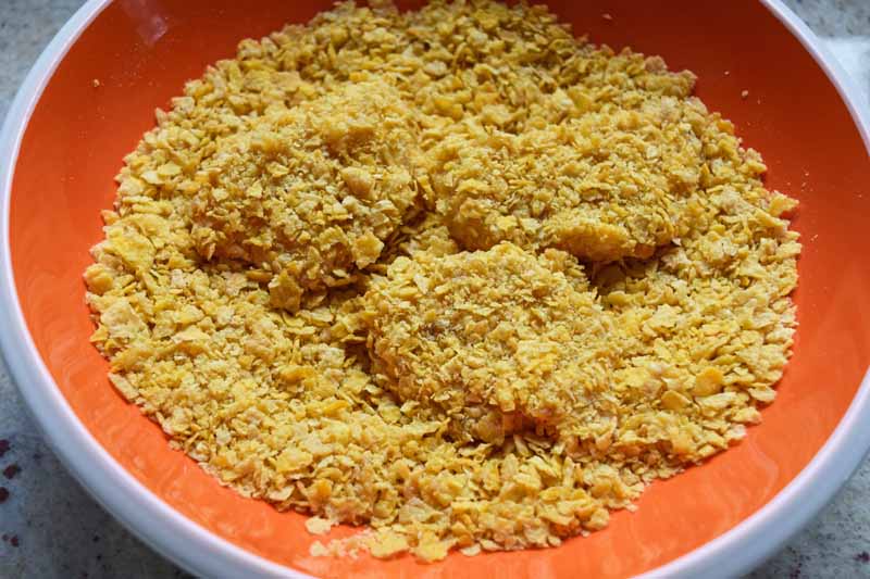 Egg covered raw chicken chunks being covered in crushed corn flakes in an orange bowl.