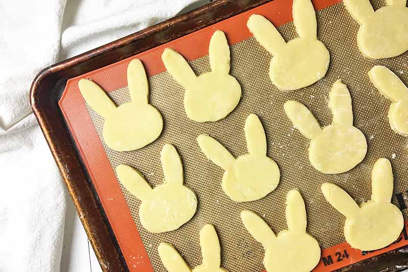 Horizontal image of unbaked rabbit cut-outs on a sheet pan lined with a silicone mat.