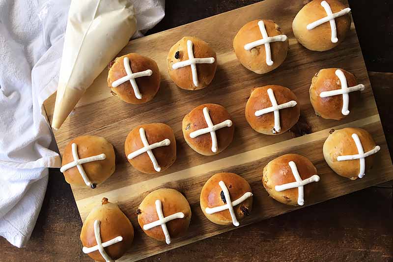 Horizontal image of decorated hot cross buns on a wooden board next to a piping bag filled with white frosting.