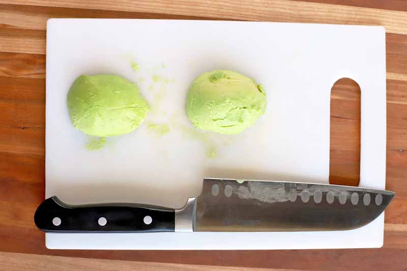 Overhead shot of two peeled halves of an avocado on a white plastic cutting board with a santoku knife with a black handle, on a wood surface.