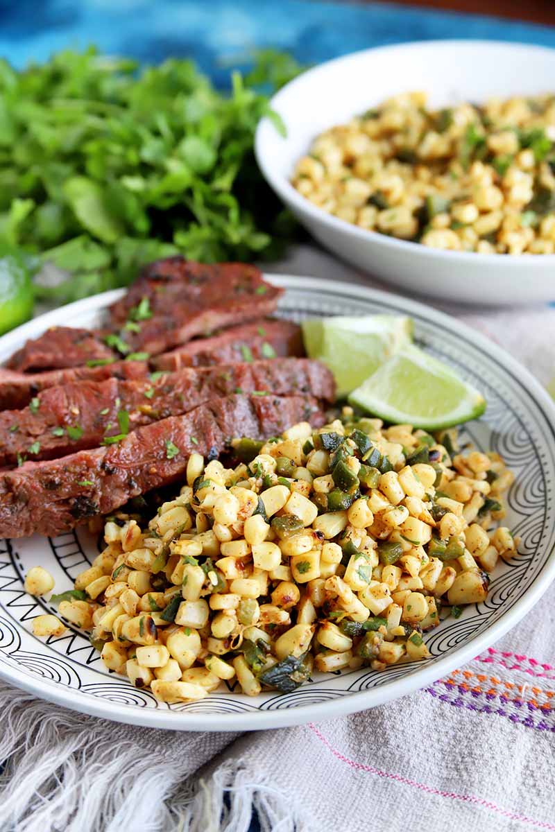 Vertical image of a plate with corn and steak and a bowl of corn next to a bunch of cilantro.