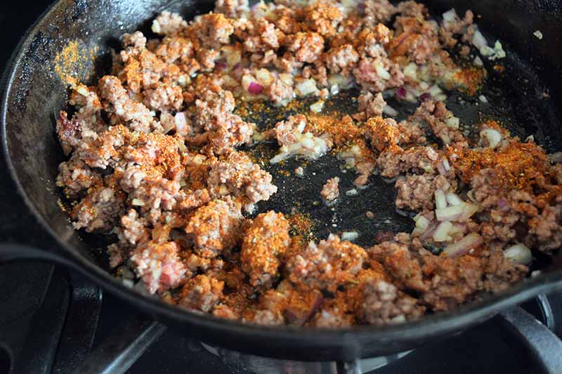 Browned ground beef cooked with onions and garlic is spread in a large frying pan, with a homemade taco spice blend sprinkled on top, on a gas stove.