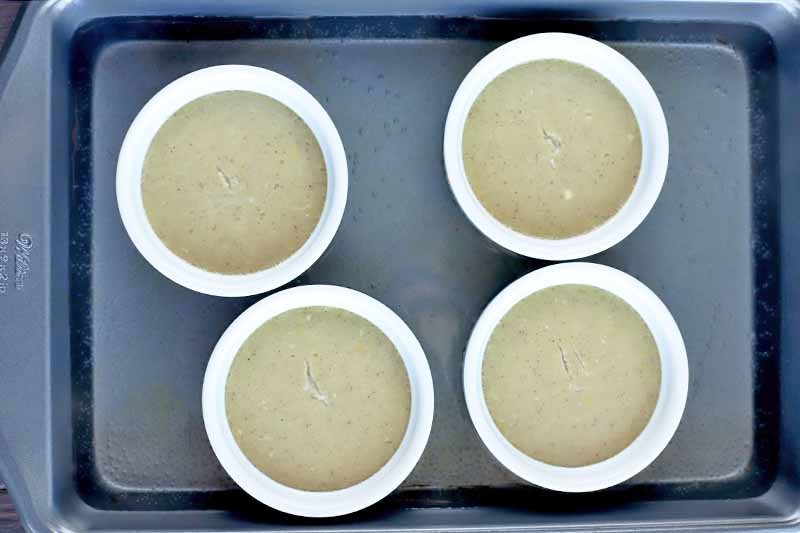Overhead horizontal shot of four white ramekins of cooked coconut custard with tops that are slightly cracked at the centers, in a metal baking pan.