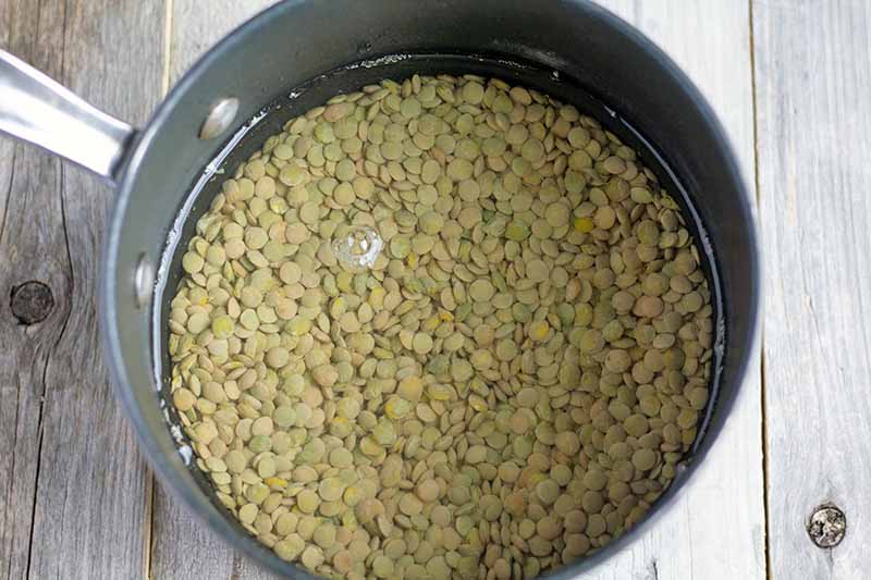 Overhead horizontal image of green lentils covered with water in a nonstick saucepan, on an unfinished wood surface.
