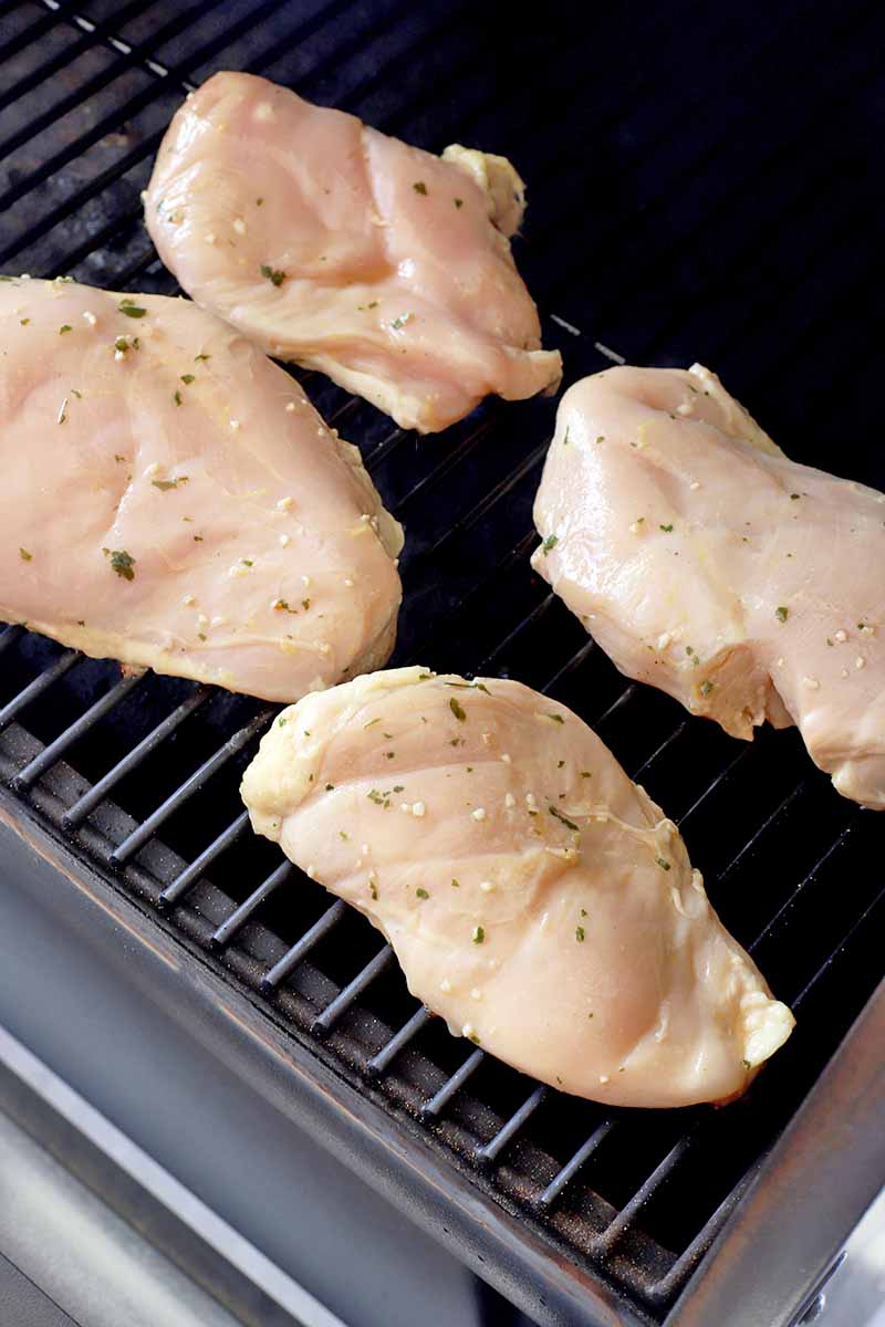 Vertical image of a grill with four boneless, skinless chicken breasts that have been coated in a marinade containing minced garlic and fresh herbs on top of the grate.