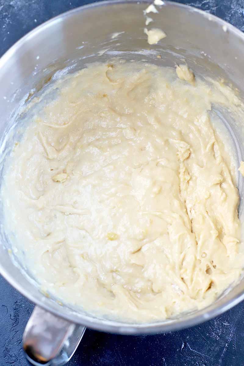 A lumpy beige batter is at the bottom of a stainless steel mixer bowl in this vertical overhead image, on a dark gray surface.