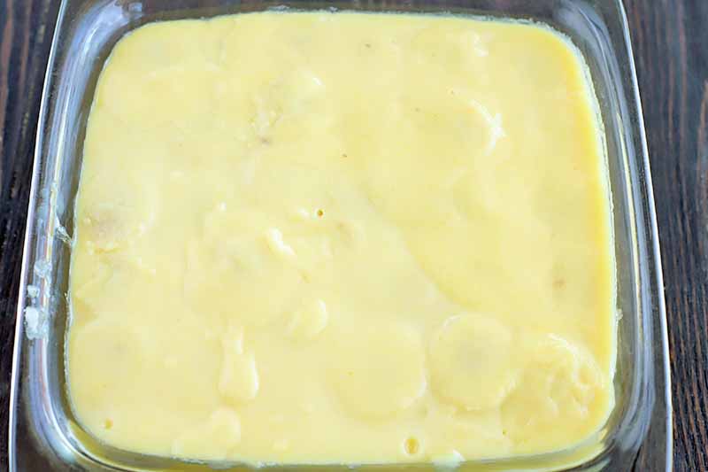 Horizontal closely cropped oblique overhead shot of a yellow pudding mixture spread mostly smoothly in a square glass baking dish, on a dark brown wood surface.