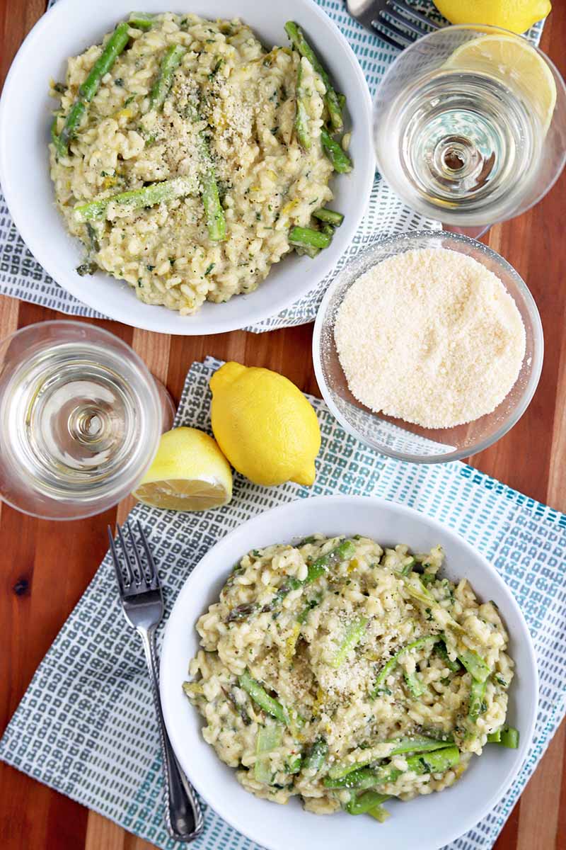 Overhead shot of two white bowls of asparagus risotto with two glasses of white wine, a small glass bowl of grated Parmigiano Reggiano, one whole and one halved yellow lemon, two light blue and white checkered cloth napkins, and a fork, on a brown wooden table.