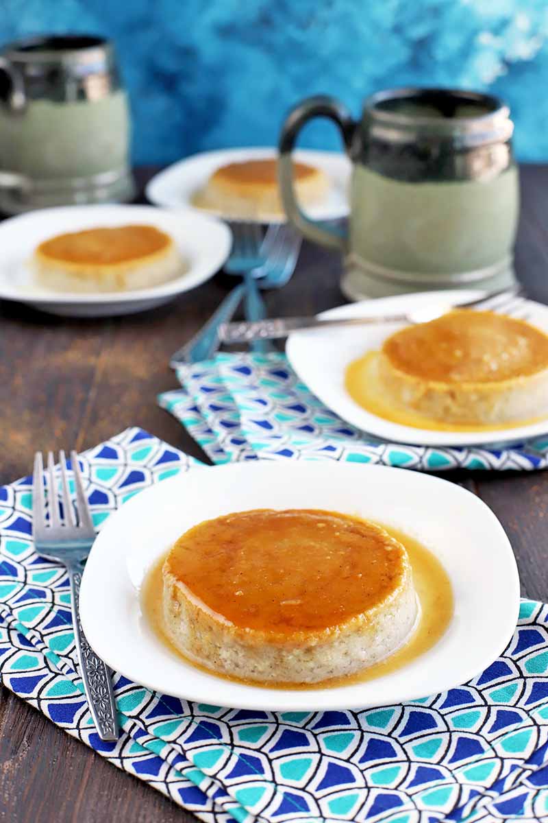 Vertical image of four white plates of coconut milk flan with caramel sauce, with two gray mugs and folded blue cloth napkins with a triangular design, and several scattered forks, on a brown wood table.