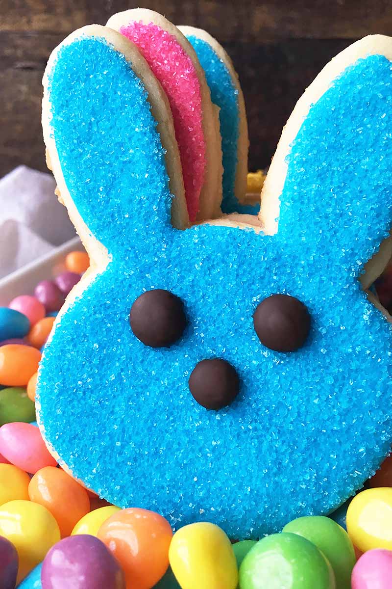 Vertical close-up image of a sugar cookie bunny with blue sprinkles on a plate with jelly beans.