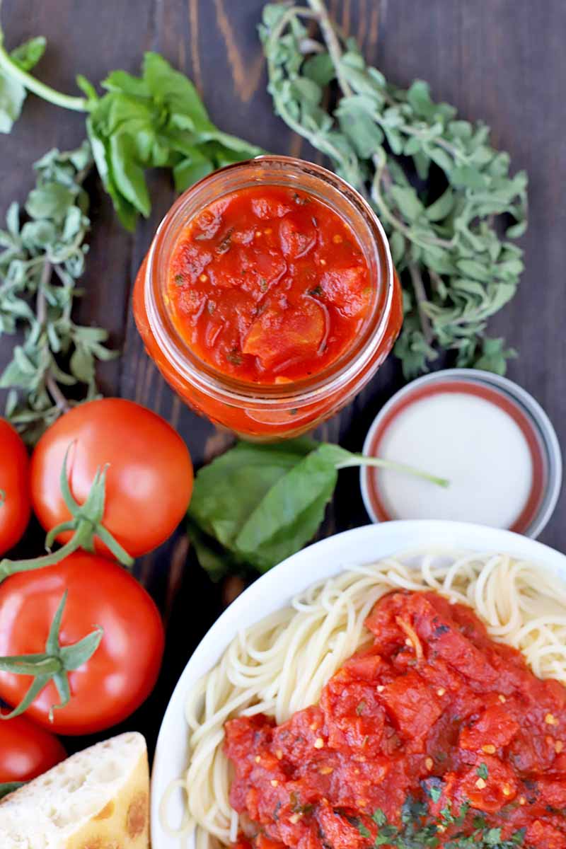 Vertical overhead image of a jar of red sauce, several red tomatoes on the vine, sprigs of basil and oregano, a metal jar lid, a piece of crusty baguette, and a white plate of cooked spaghetti topped iwth chunky marinara and a garnish of fresh herbs, on a dark brown wood surface.