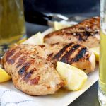 Horizontal image of four grilled chicken breasts on a white serving platter with wedges of lemon, with two mugs of beer to the right and left, and a white cloth with blue stripes in the foreground, on a gray slate surface.